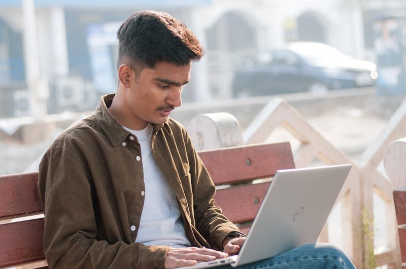 A young man focused on his laptop, working outdoors on a sunny day.