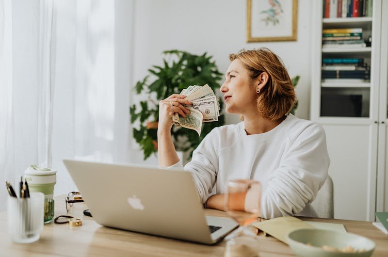 Woman sitting at desk holding cash while working on a laptop in a cozy home office setting.